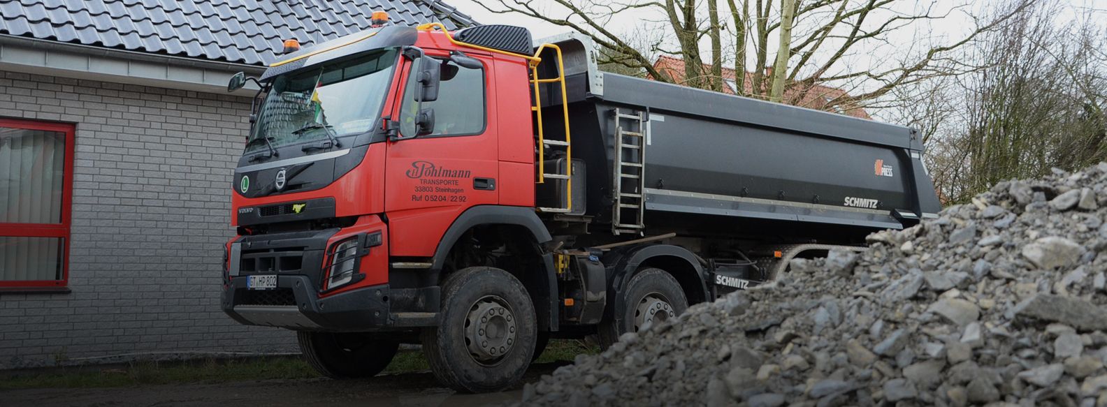 Rot-schwarzer Transporter der Firma Pohlmann zwischen einem Haus und einem Berg Schüttgut